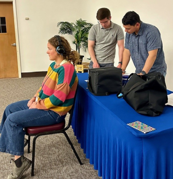 Two grad students working with equipment while client sits with headphones on in front of them